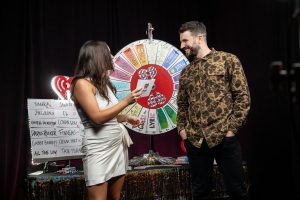 Curl holds up an iHeart radio card next to singer Sam Hunt in front of a spinning wheel for part of a game show event