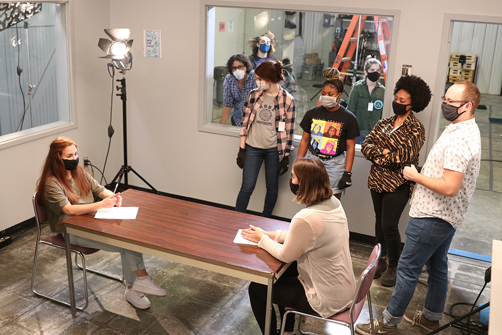 Two ladies at a table and four students standing around with light in the corner, ready to film a scene.
