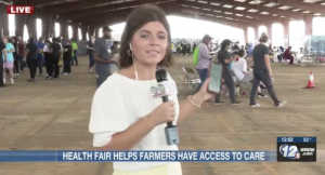 Sydney doing a liveshot on camera, holding a microphone and pointing to a group of people at a health fair