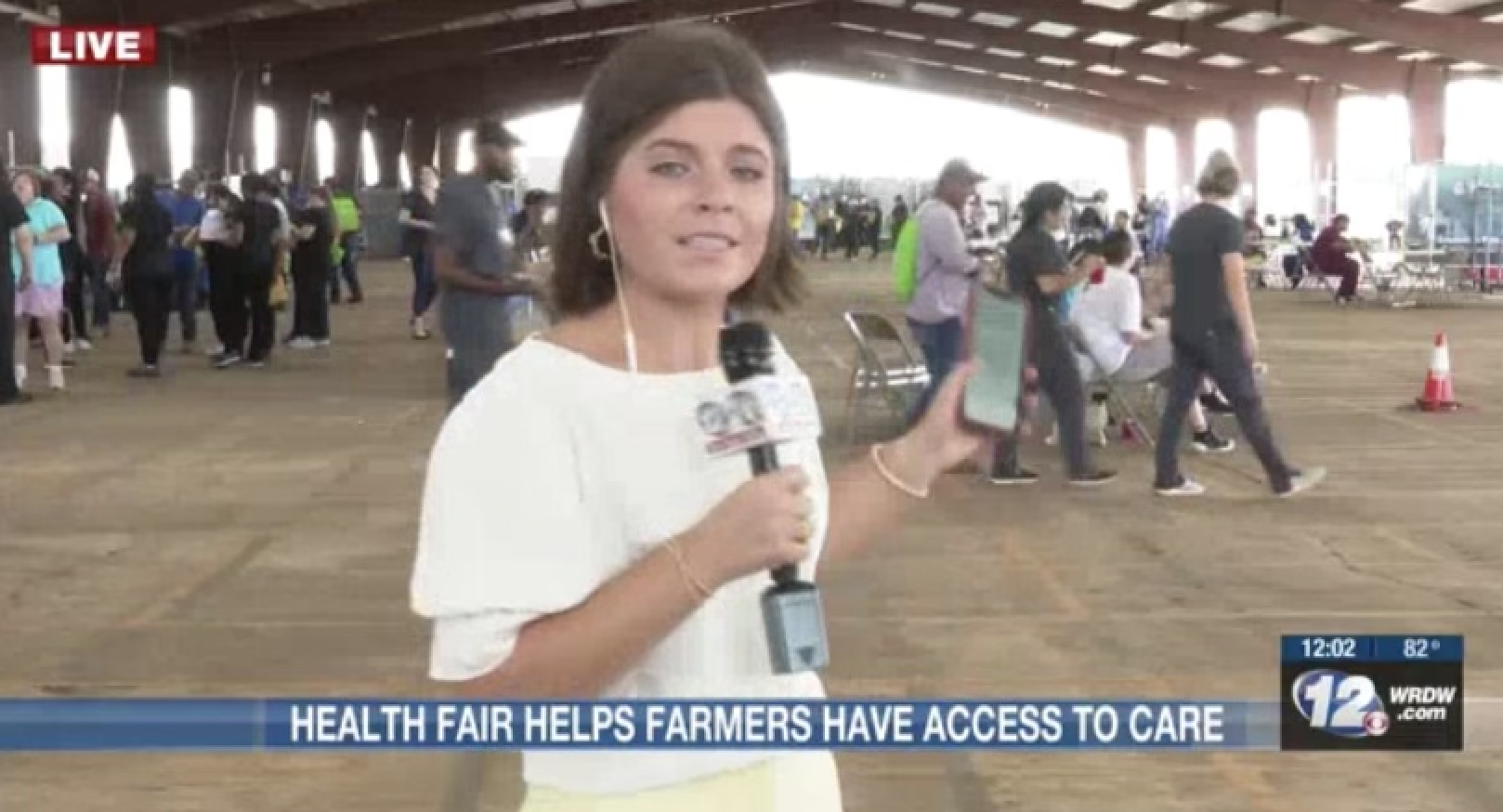 Sydney doing a liveshot on camera, holding a microphone and pointing to a group of people at a health fair