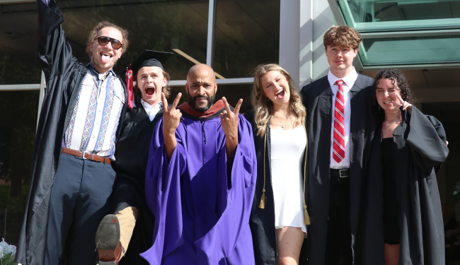 Booker Mattison, wearing a purple graduation robe, holds up backwards peace signs, surrounded by students.