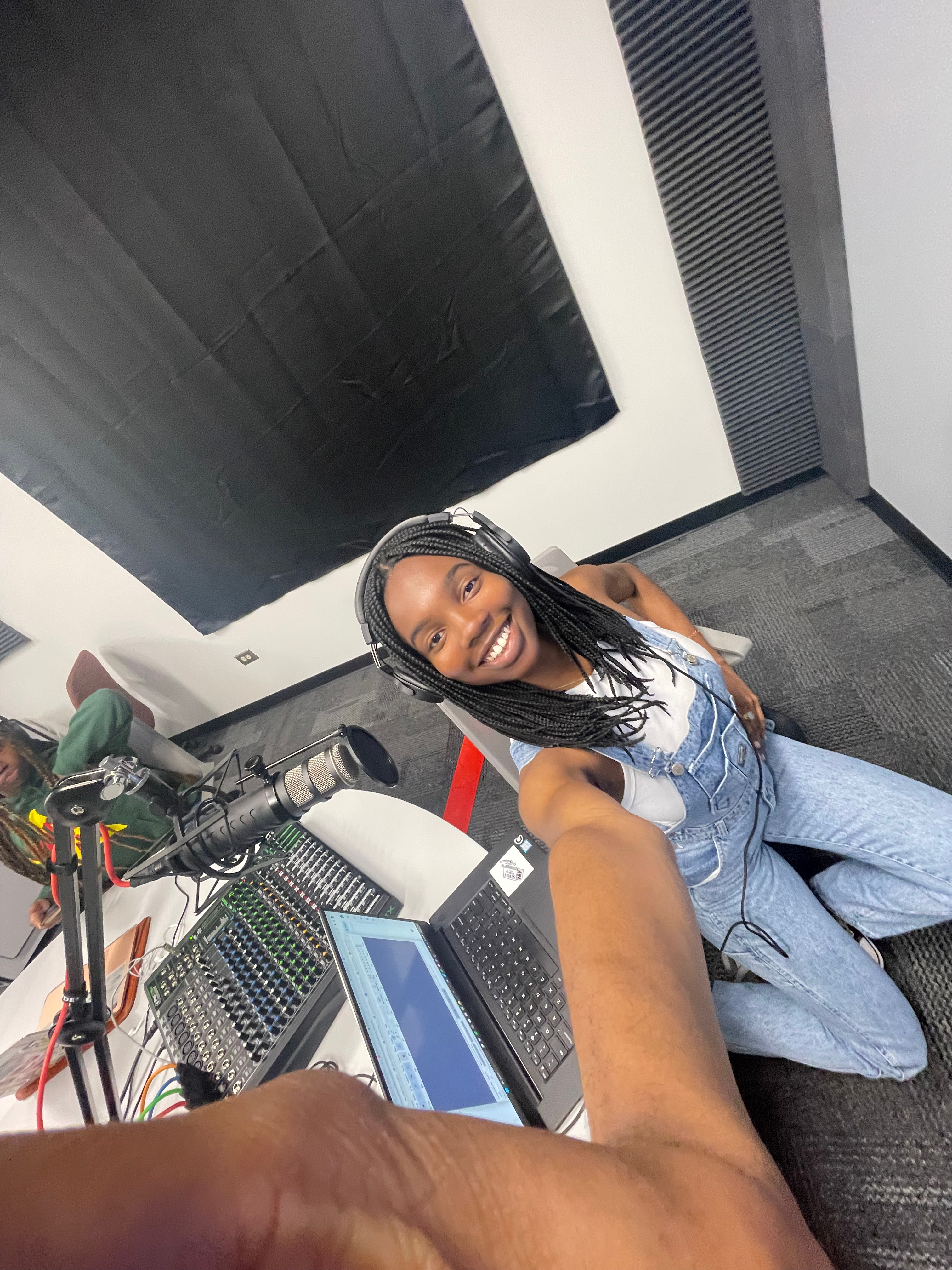 Chiamaka Uwagerikpe sitting in a chair in a podcast studio holding up her phone to take a selfie. 