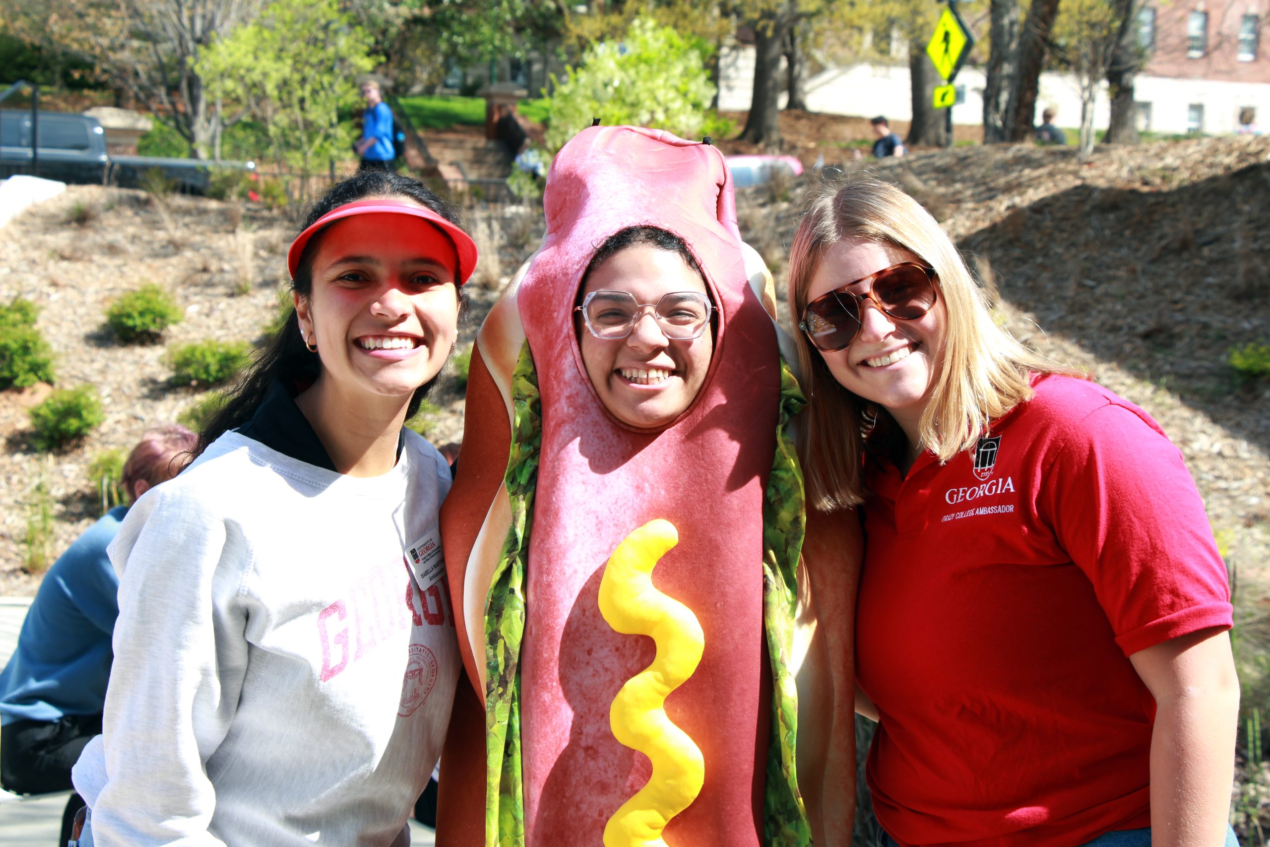 Three students, including one in the middle with a hot dog costume on, pose for a photo outside during Dawgs with the Dean.