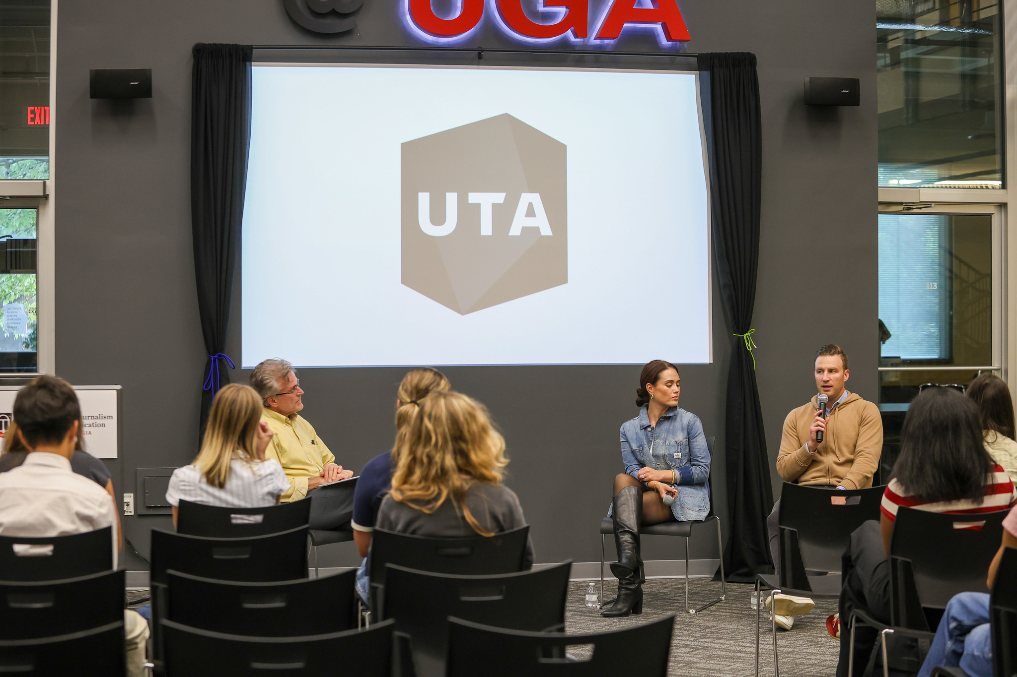 A panel discussion with two professionals in front of a group of students. In the background is a digital screen reading UTA.