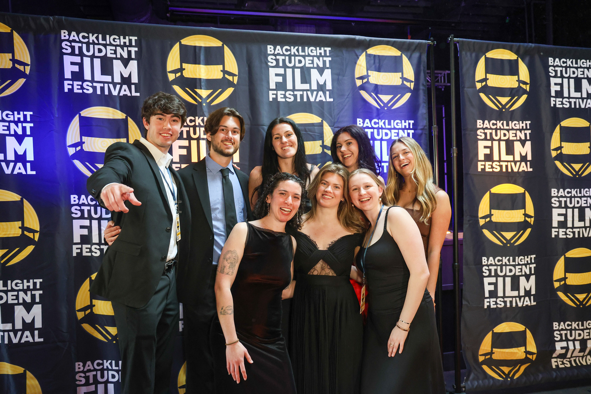 A group of students and alumni in formal attire pose for a picture in front of a Backlight Film Festival backdrop.