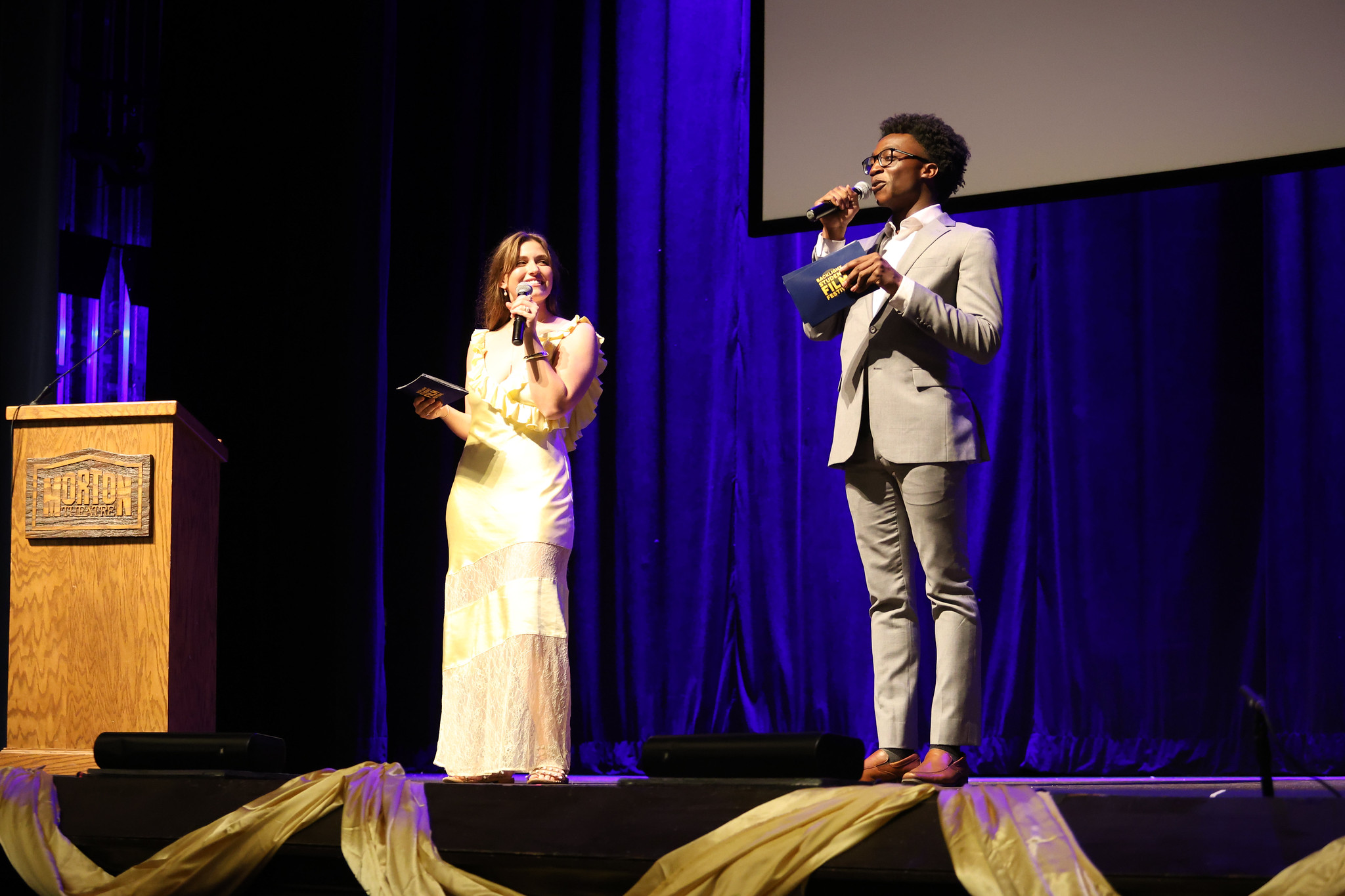 Two students in formal attire address a group from a stage.