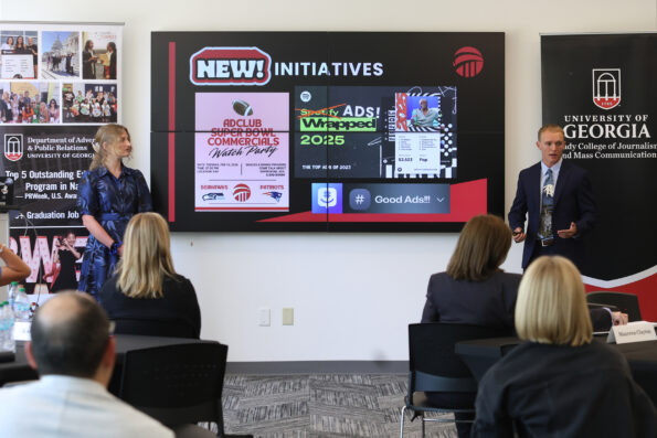 A male and female student make a presentation to a group of people. In the background