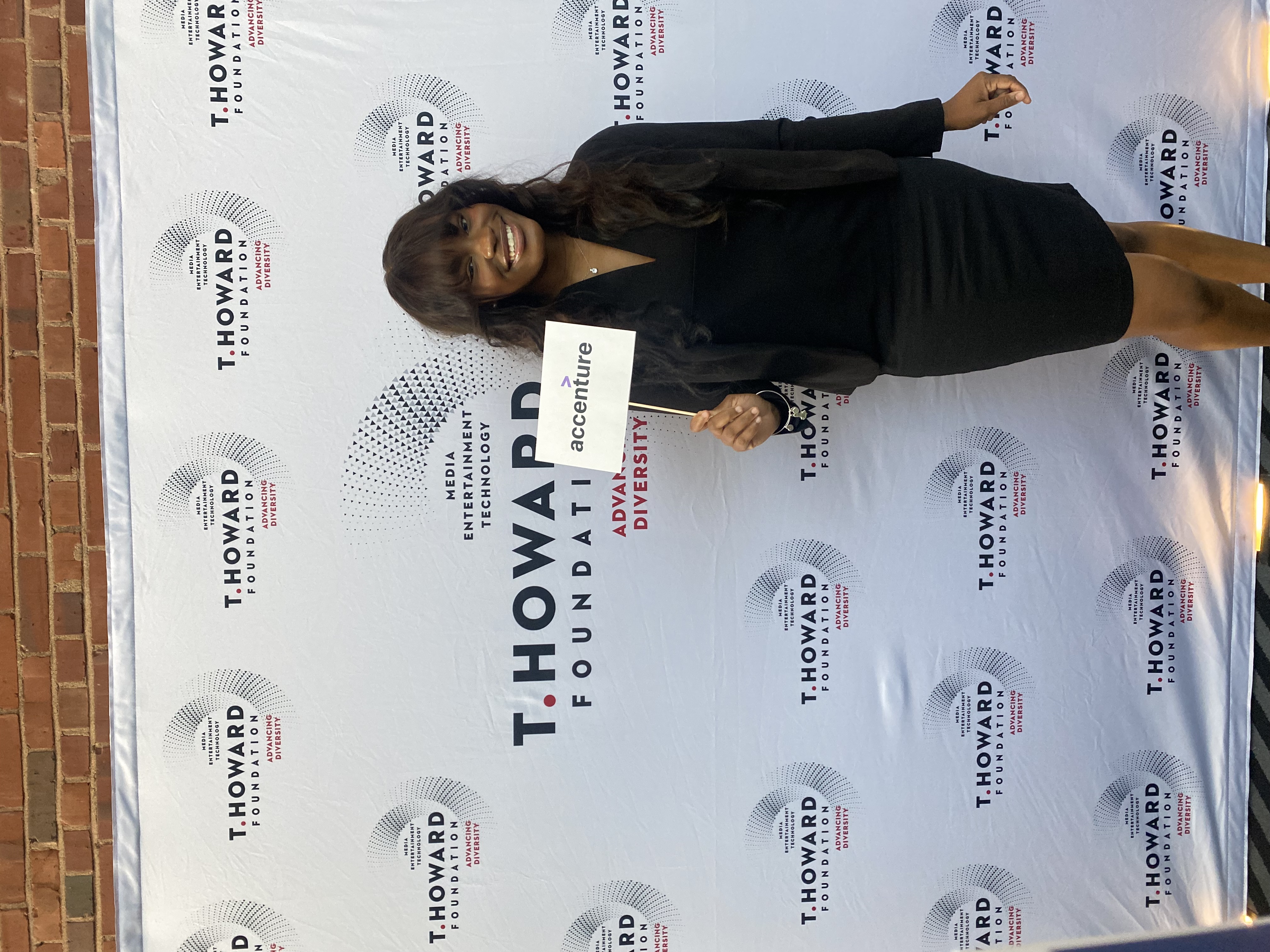 Geter stands in front of a backdrop with the text T. Howard Foundation on it. She holds a sign that says Accenture.