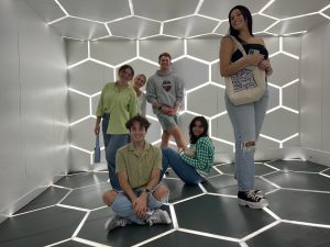 5 students pose for a picture in a room lit up with glowing pentagon shapes on the floor, ceiling and walls