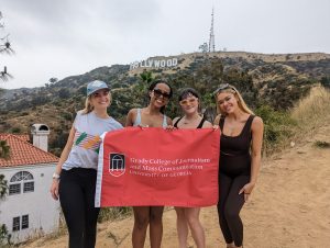 Four students holding a Grady College flag pose in front of the Hollywood sign