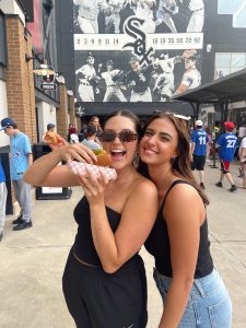 Two students pose for a picture while eating a hot dog in a baseball stadium