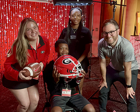 Boy holds a UGA football helmet while football staff and another student pose nearby.