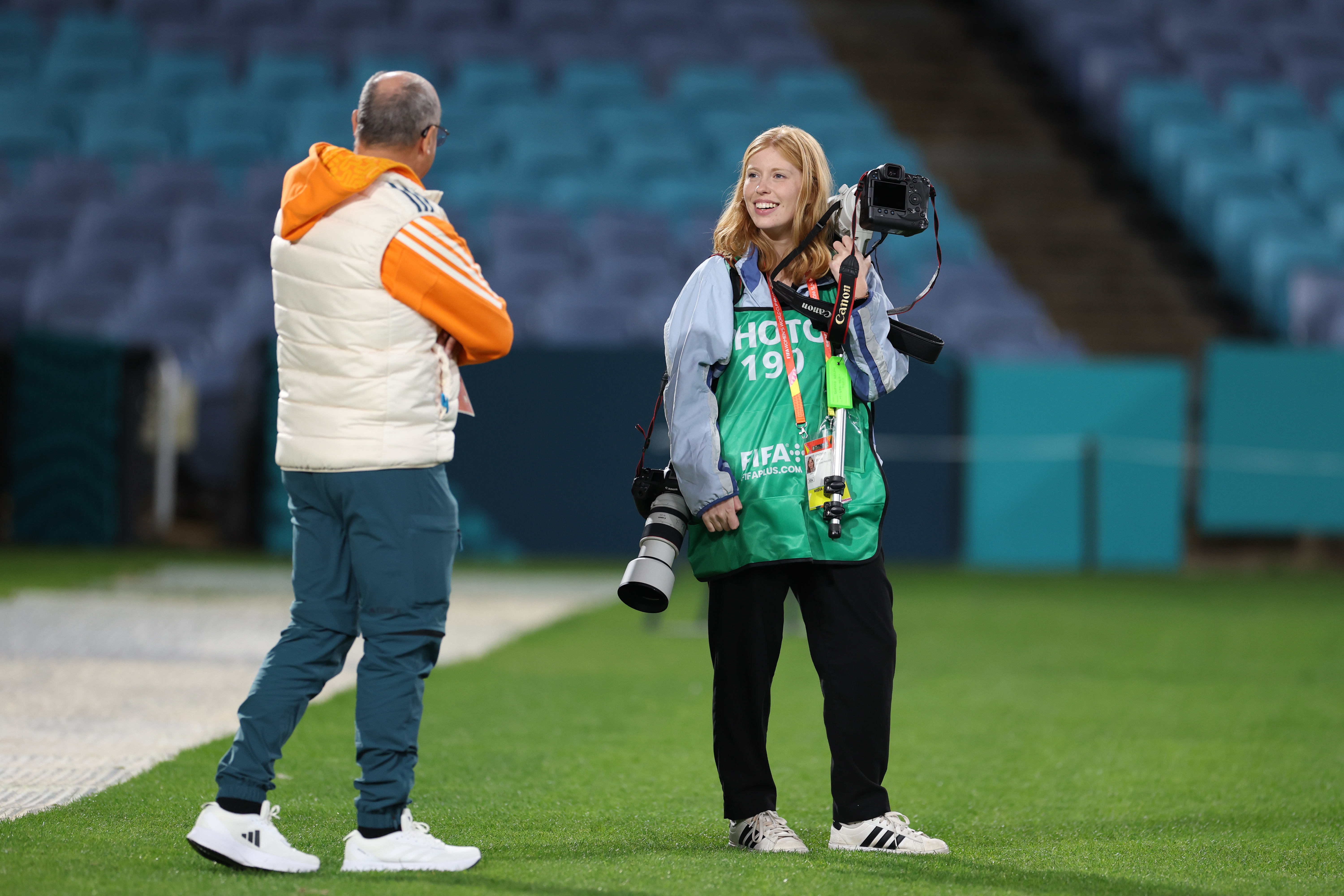A student photographer stands with with two cameras on a soccer field talking with a coach. 