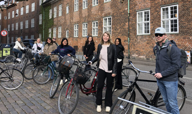 Students stand in a line with bicycles and a large brick building behind.