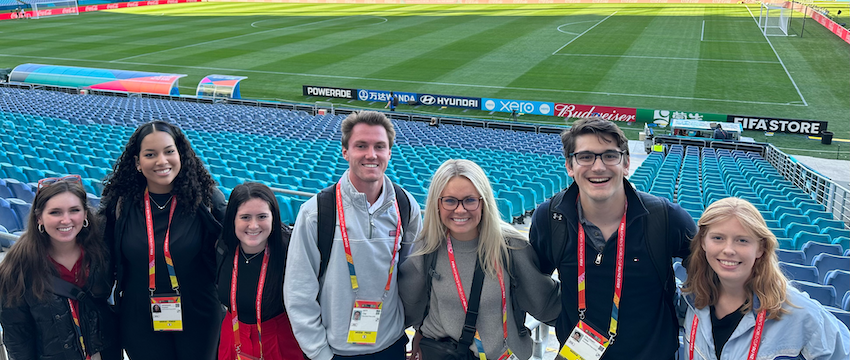 Group of seven students pose for a photo in front of and empty soccer stadium at the Women's World Cup in Australia.