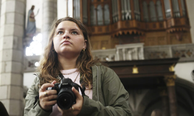 A close-up of a female student holding a camera and looking in the distance. Behind her are the pipes of a large church organ in a Prague cathedral.