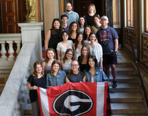 A large group of students and professors stand on stairs and the front row is holding a UGA flag. 