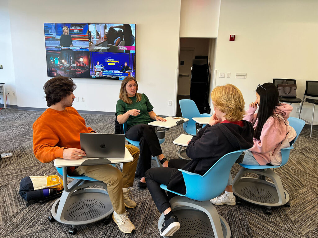 Four people sit in a circle on blue chairs, engaged in discussion. One woman speaks while three students from the Cox Institute listen, taking notes or using a laptop. Multiple TV screens with news are visible on the wall behind them.