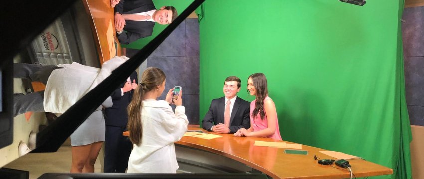 Two students sit in front of a green screen as they prepare to film a segment.