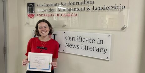 A smiling young woman in a red Georgia shirt holds a Certificate of Achievement in front of a wall sign reading Certificate in News Literacy at the University of Georgias Cox Institute for Journalism.
