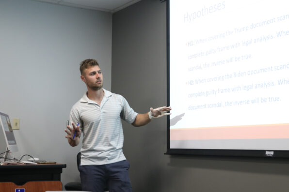 A man in a striped polo shirt stands near a projector screen, presenting and gesturing with his hands. The screen displays text titled Hypotheses in a classroom or conference room setting.