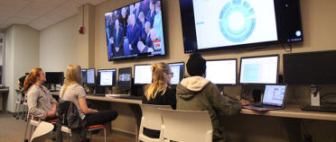 Students look at computer screens with large displays monitors overhead.