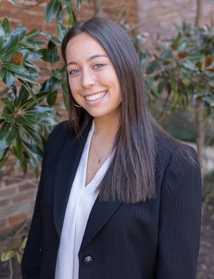 A young woman with straight brown hair, wearing a white blouse and a black blazer, smiles while standing outdoors in front of green leafy plants and a brick wall, representing the Grady College Ambassadors.