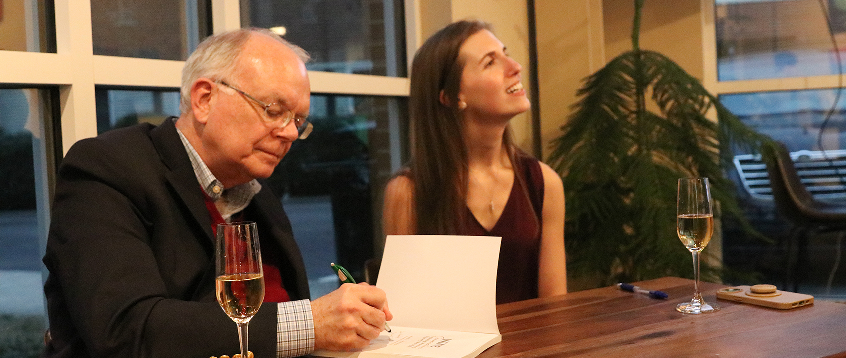 Keith signs a book at a table while Charlotte smiles and talks with someone out of frame.