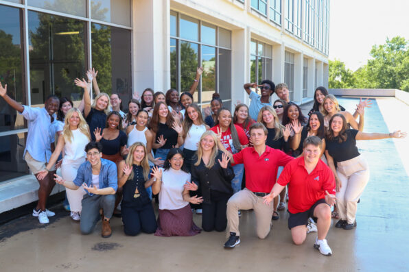 A large, diverse group of young adults pose and smile excitedly with arms raised outside a modern building with large windows on a sunny day during the Ambassador Retreat.