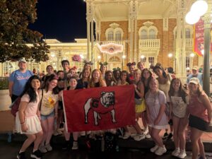 A group of students, mostly female with some male, stand in front of an old-looking building. Some of them are wearing Micky Mouse ears, most are not. There six young women in the center holding a flag with a University of Georgia Bulldog on it. 