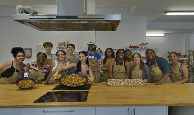 A group of about 13 students in a large industrial kitchen pose for a picture. Two of them are holding dishes they prepared: a pizza and a casserole.