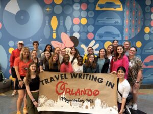 A large group holds up a banner that reads "travel writing in Orlando Maymester 2026."