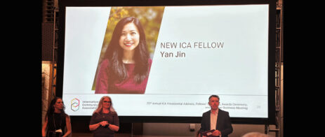 People stand in front of a projector screen with a picture of Yan Jin and the worlds "New ICA Fellow, Yan Jin."
