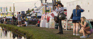 Photographers along a waterway in the foreground look in their viewfinders with fair rids and tents in the background.