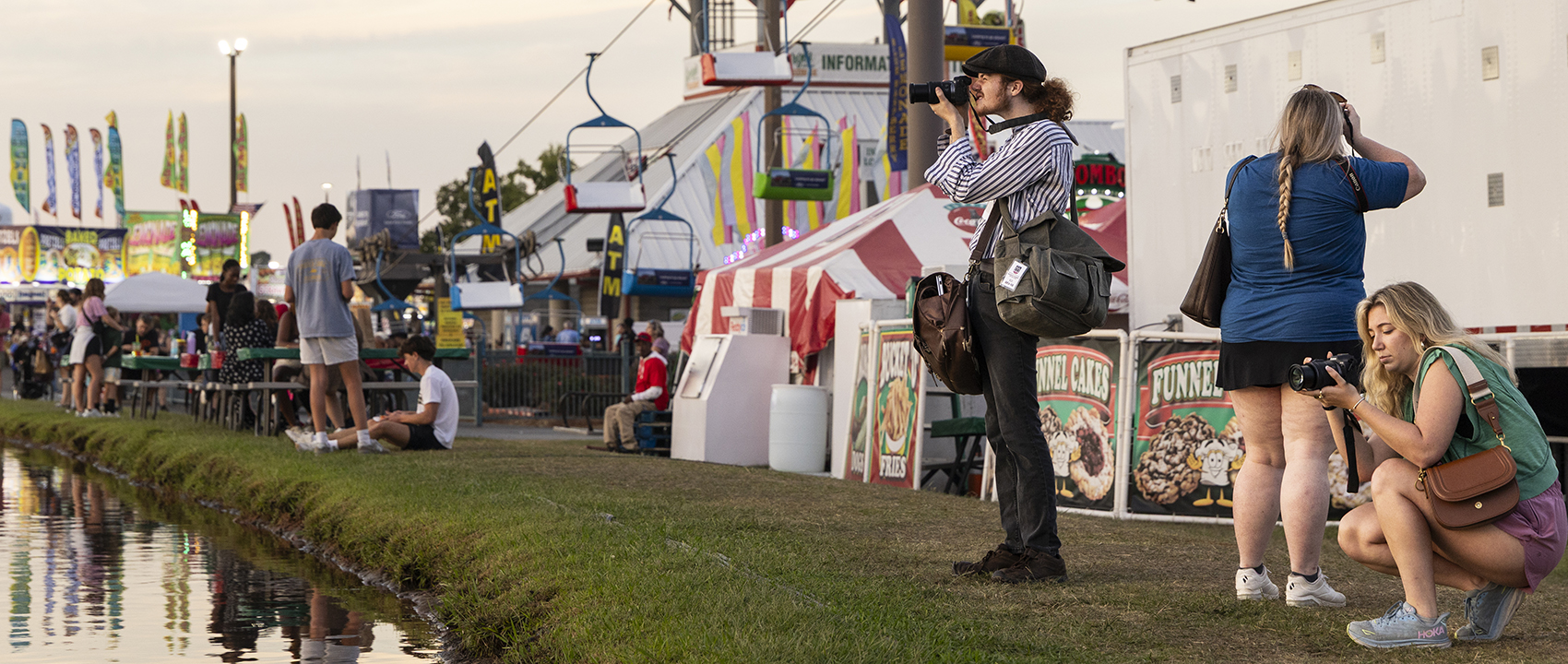 Photographers along a waterway in the foreground look in their viewfinders with fair rids and tents in the background.