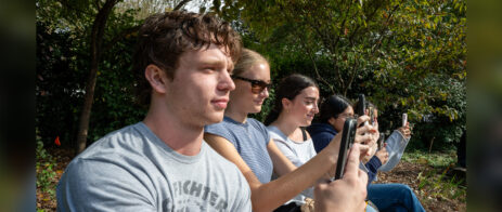 Students sit on a bench in a line as they hold up their phones to record.