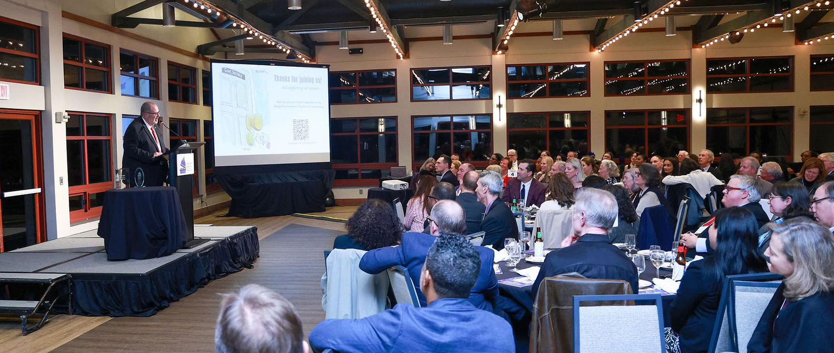 Charles Davis stands at a lectern and addresses a group of people at tables.
