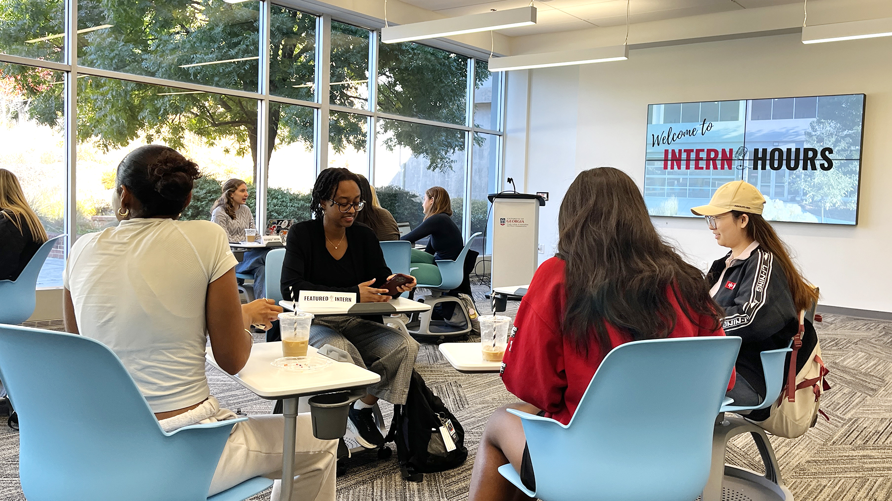 Group of students sit in a circle talking while a screen says Welcome to Intern Hours in the background.