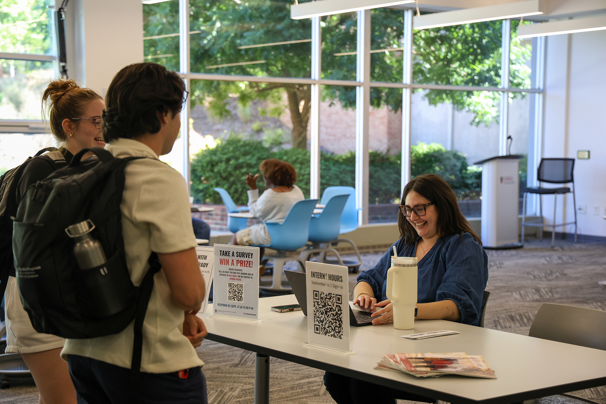 Woman sitting at a table laughs while registering students for an event. A sign that reads Intern Hours sits on the table.