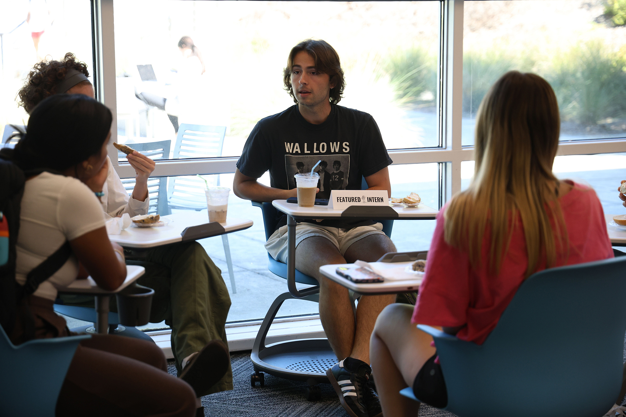Male student sits in a chair and talks with other students. A sign in front of the student reads Featured Intern.