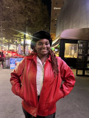 A smiling person in a red leather jacket, white shirt, and black hat stands outdoors at night—an inspiring figure among the Profiles of Tenacity—with trees, lights, and city buildings in the background.