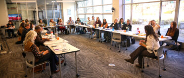 An overhead view of a woman talking with a group of around 30 students sitting at u-shaped tables.