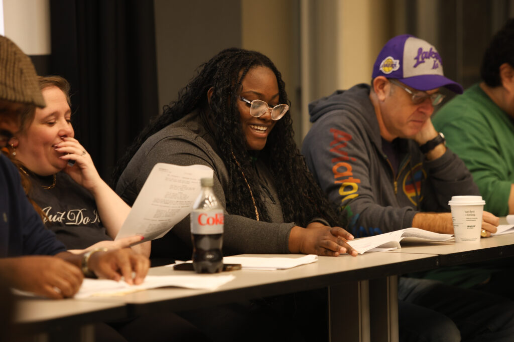 A woman smiles as she sits at a table and reads a script.
