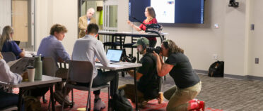 A film crew records some students working on their laptops at a table.