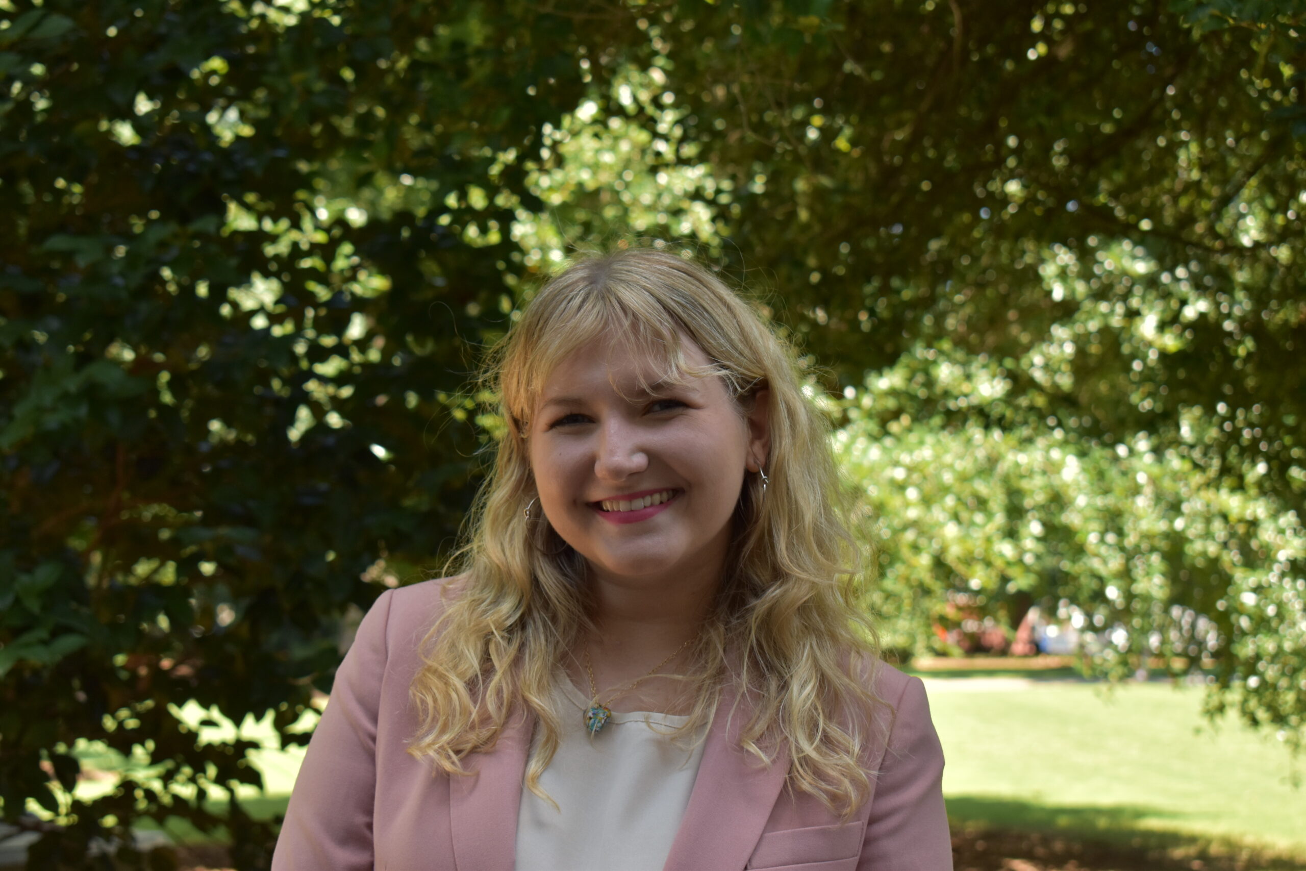 A young woman with long blonde hair, wearing a pink blazer and white top, smiles outdoors in a sunny, green park with trees in the background.
