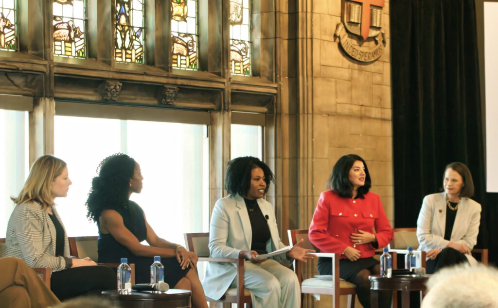 Five women, including Tracy Brown, sit on a panel discussion in front of stained glass windows and a stone wall with a crest, each dressed in business attire and engaging in conversation with water bottles on the table.