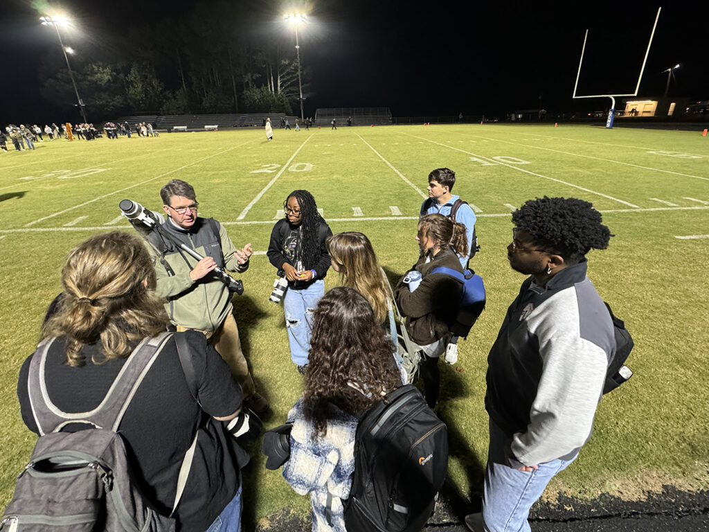 Mark Johnson stands in a group of students off to the side of a football field explaining something.