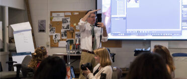 Mark Johnson stands in front of a classroom and gestures to show space between his hands as students look on.