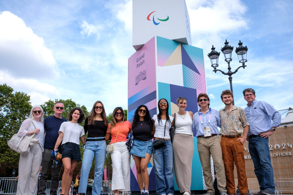 Mark Johnson and Welch Suggs pose for a picture with a group of students standing in front of a Paralympics signage that reads "Grand Palais."