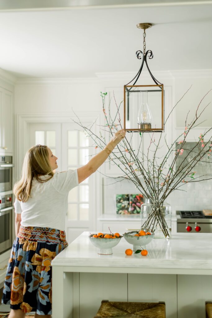 Robin Burnett is pictured in a space designed by her company, Robin Burnett Design. She is reaching up to straighten a lamp above an island in a kitchen.